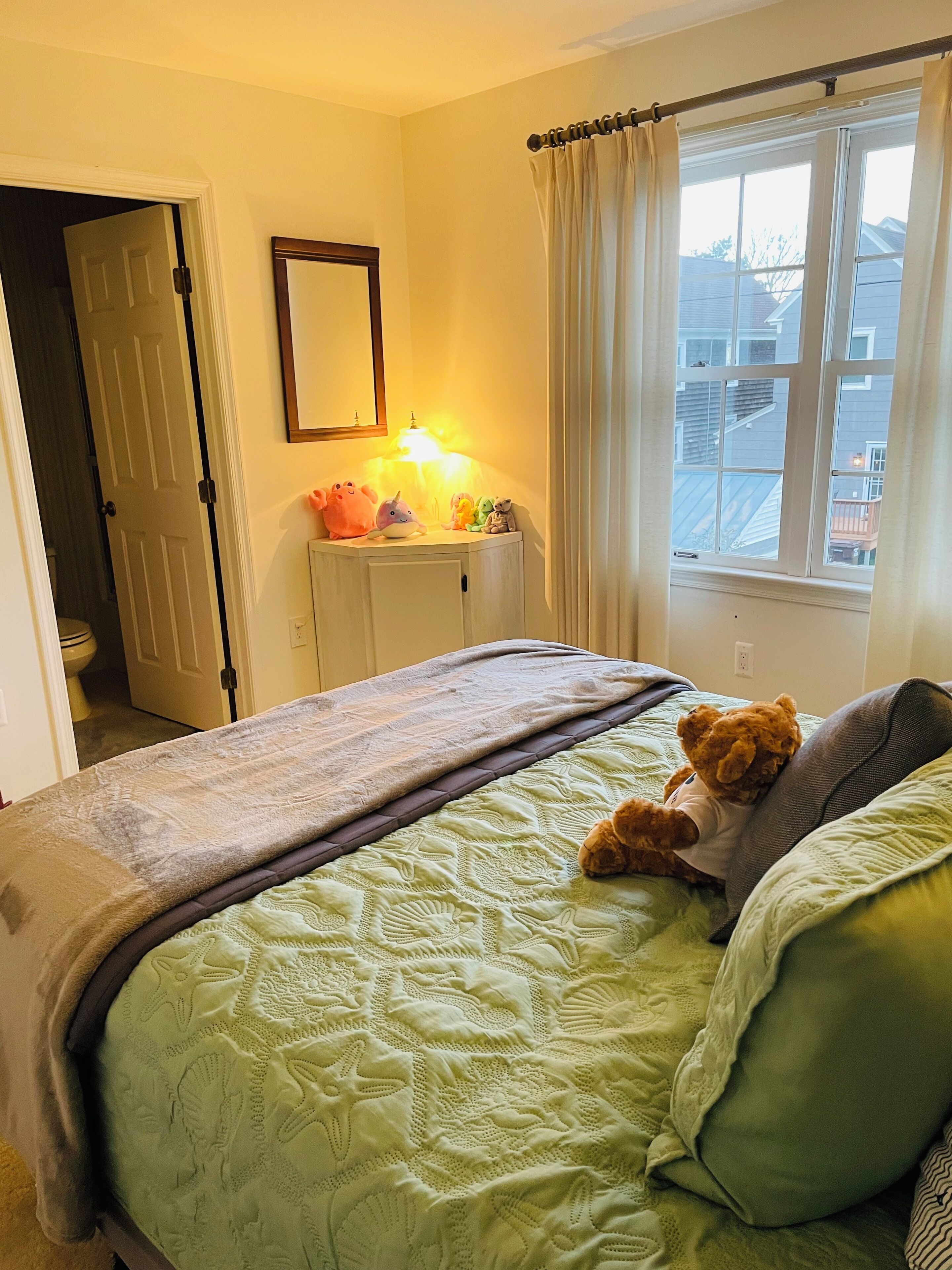A bedroom with a bed, a bedside table with plush toys, a window with curtains, and a door leading to another room.