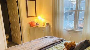 A bedroom with a bed, a bedside table with plush toys, a window with curtains, and a door leading to another room.