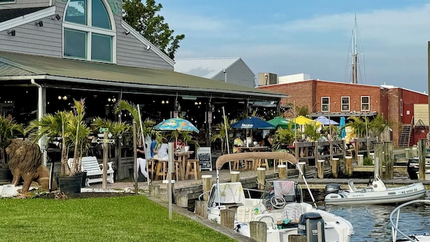 Un restaurant situé au bord de l’eau, doté d’une terrasse, d’un quai avec des bateaux et d’un bâtiment surmonté d’une marquise verte.