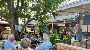 Un espace de restauration en plein air, avec des tables et des chaises en bois, entouré de verdure et d’un grand arbre.