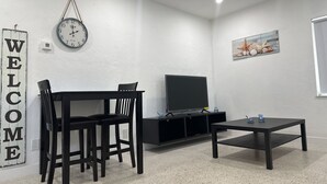 A modern living room with a black TV stand, a coffee table, and a dining table with chairs. A clock and a 'WELCOME' sign are on the wall. - Beach Apartment in Hollywood  (Hollywood)