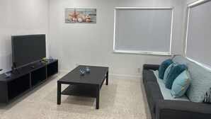 A living room with a flat-screen TV, a coffee table, a sofa with cushions, a painting on the wall, and a window with blinds. - Beach Apartment in Hollywood  (Hollywood)