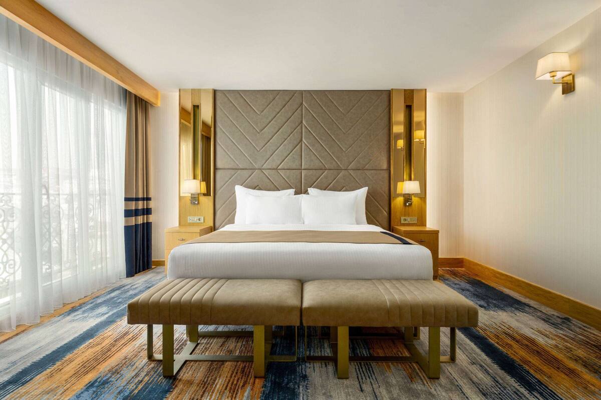 Une chambre d’hôtel avec un grand lit, deux tables de chevet, un banc, une vue sur un balcon et une tête de lit décorative.