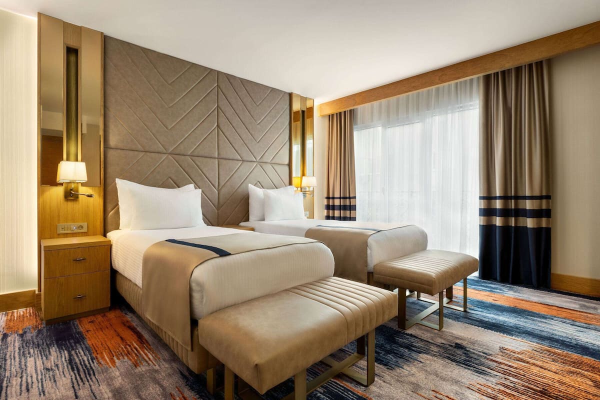 Une chambre d’hôtel avec deux lits, une banquette, une table de chevet et une grande fenêtre avec des rideaux.