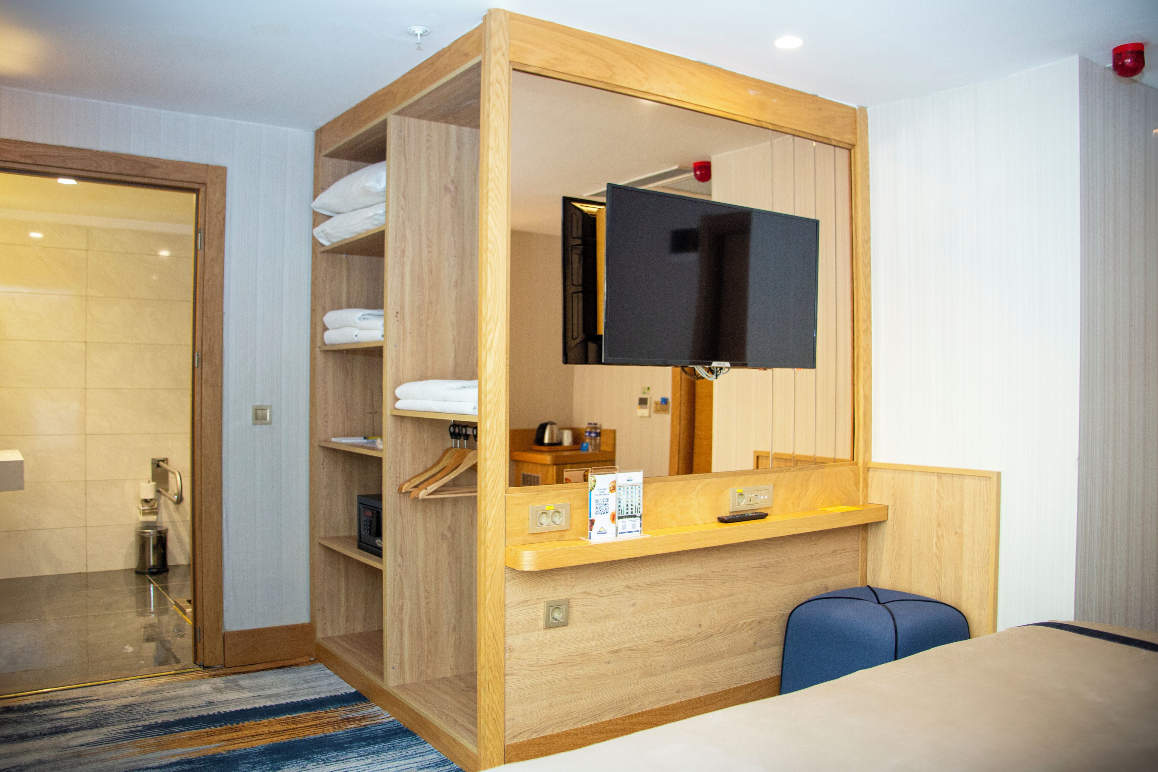 A hotel room with a wooden wardrobe, a television mounted on the wall, a small desk, and a blue chair.