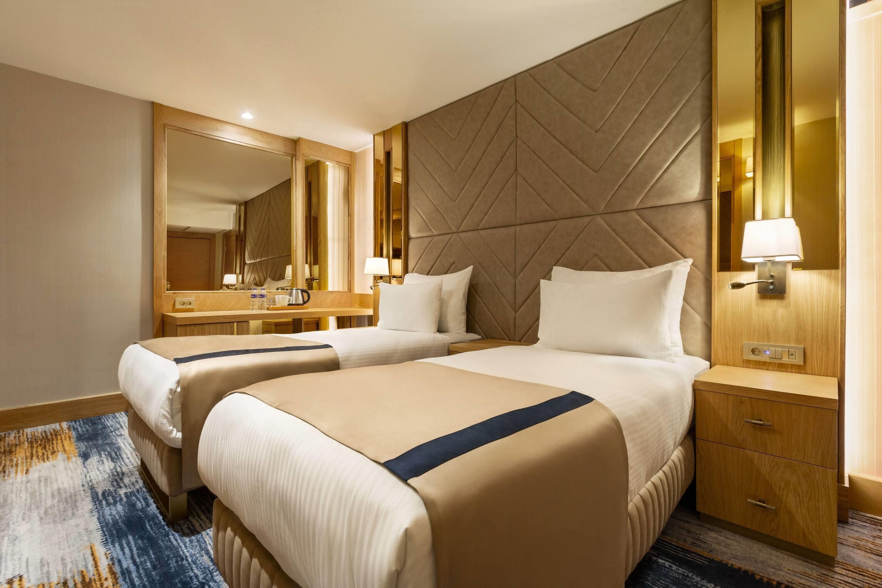 A hotel room with two beds, a large mirror, a bedside table with a lamp, and a wooden nightstand.