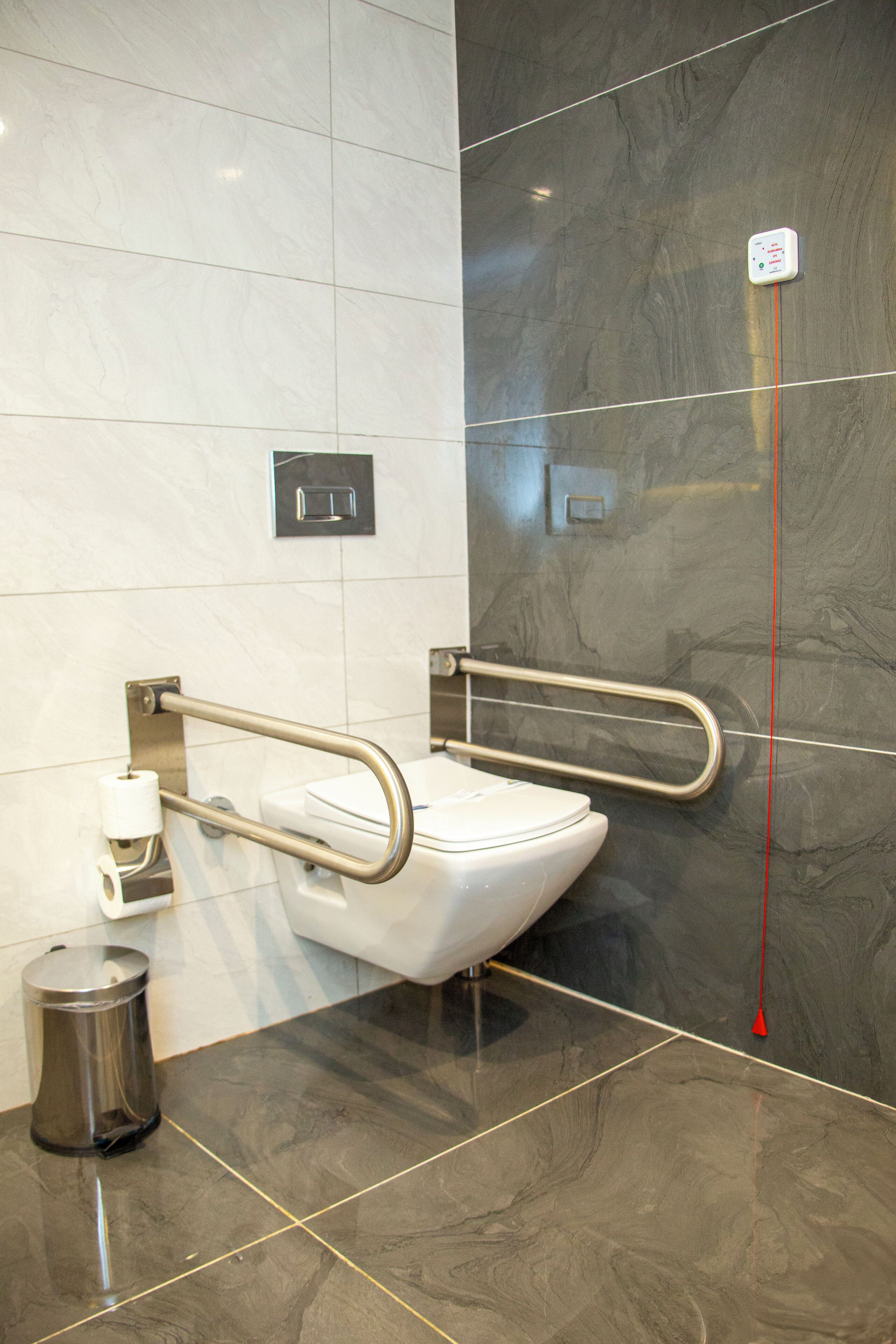 Un baño moderno con inodoro blanco, barra de apoyo, cubo de basura y pulsador de descarga en la pared.