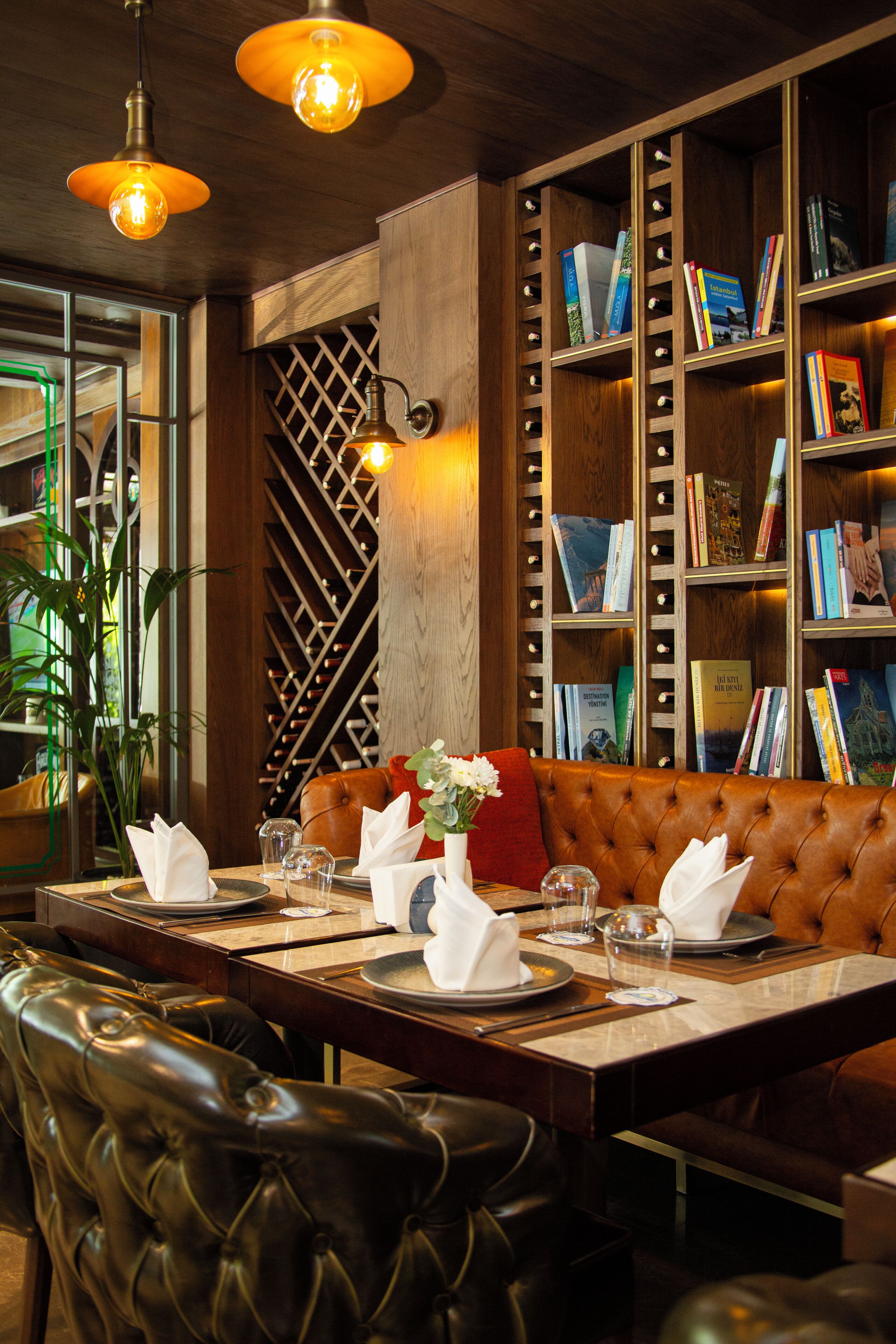 A restaurant with a long wooden table set for dining, cushioned seating, and a bookshelf filled with books.