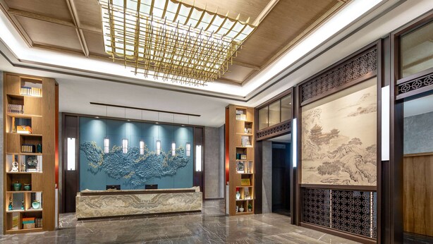 A reception area with a marble counter, wooden shelving with decorative items, a large wall mural, and a ceiling with a geometric light fixture.