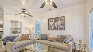 A modern living room with two sofas, a glass coffee table, a ceiling fan, a world map artwork, and a kitchen area visible through an open door. - Centrally located to Charleston’s best! (North Charleston)