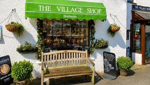 Botiga anomenada 'The Village Shop' amb tendal verd, una banca de fusta al davant i plantes penjants.