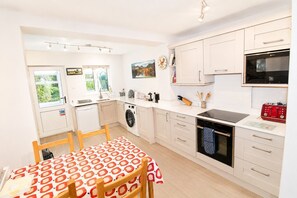 A modern kitchen with a washing machine, oven, and dining area with a table set for two. - Hill Cottage, Braithwaite with views to Skiddaw and walks from the door (Braithwaite)