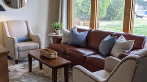 A living room with a brown leather sofa, a beige armchair, a round mirror, and a view of a green landscape through large windows. - Wingate - Meridian, Cozy Acreage in the heart of the city (Meridian)