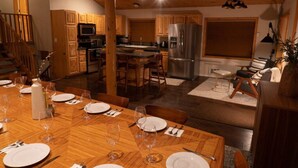 Un espace repas avec une table en bois dressée pour six personnes, une cuisine dotée de meubles en bois et d’appareils électroménagers en acier inoxydable, et un escalier menant à un niveau supérieur.