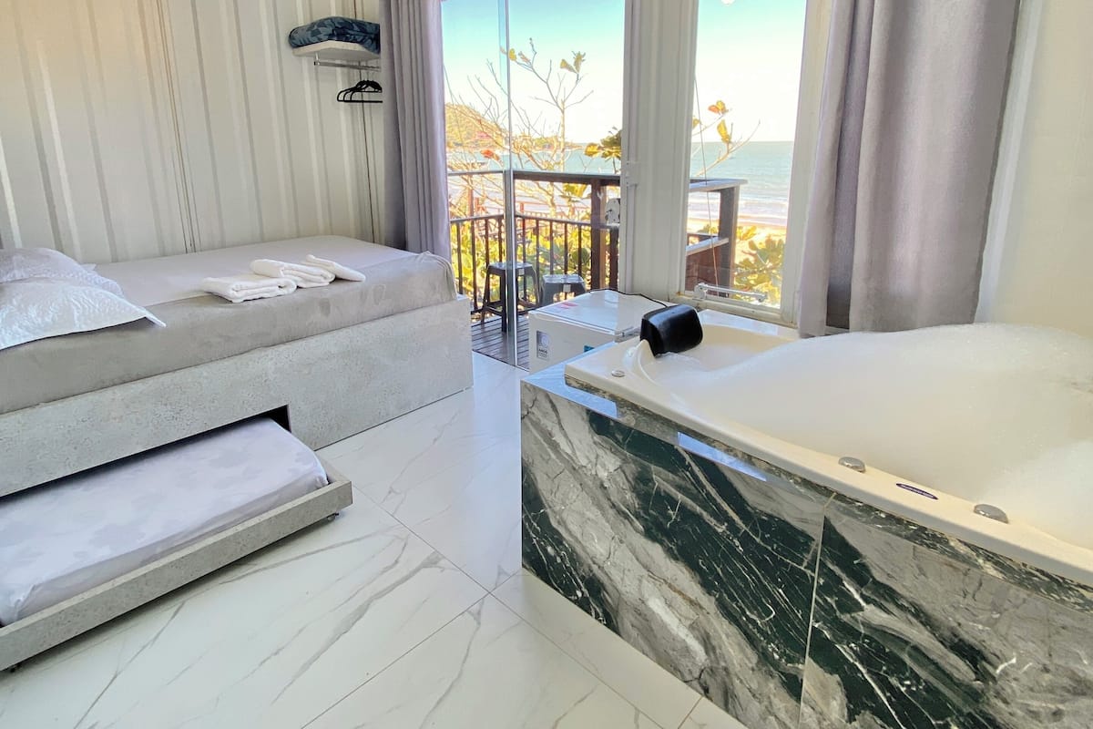 A modern bedroom with a large bed, a marble bathtub, and a balcony with a view of the sea.