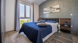 A bedroom with a large bed, a headboard, a bedside table, a nightstand, a window with curtains, a painting on the wall, and a view of greenery outside. - Villa InSa (Pican)