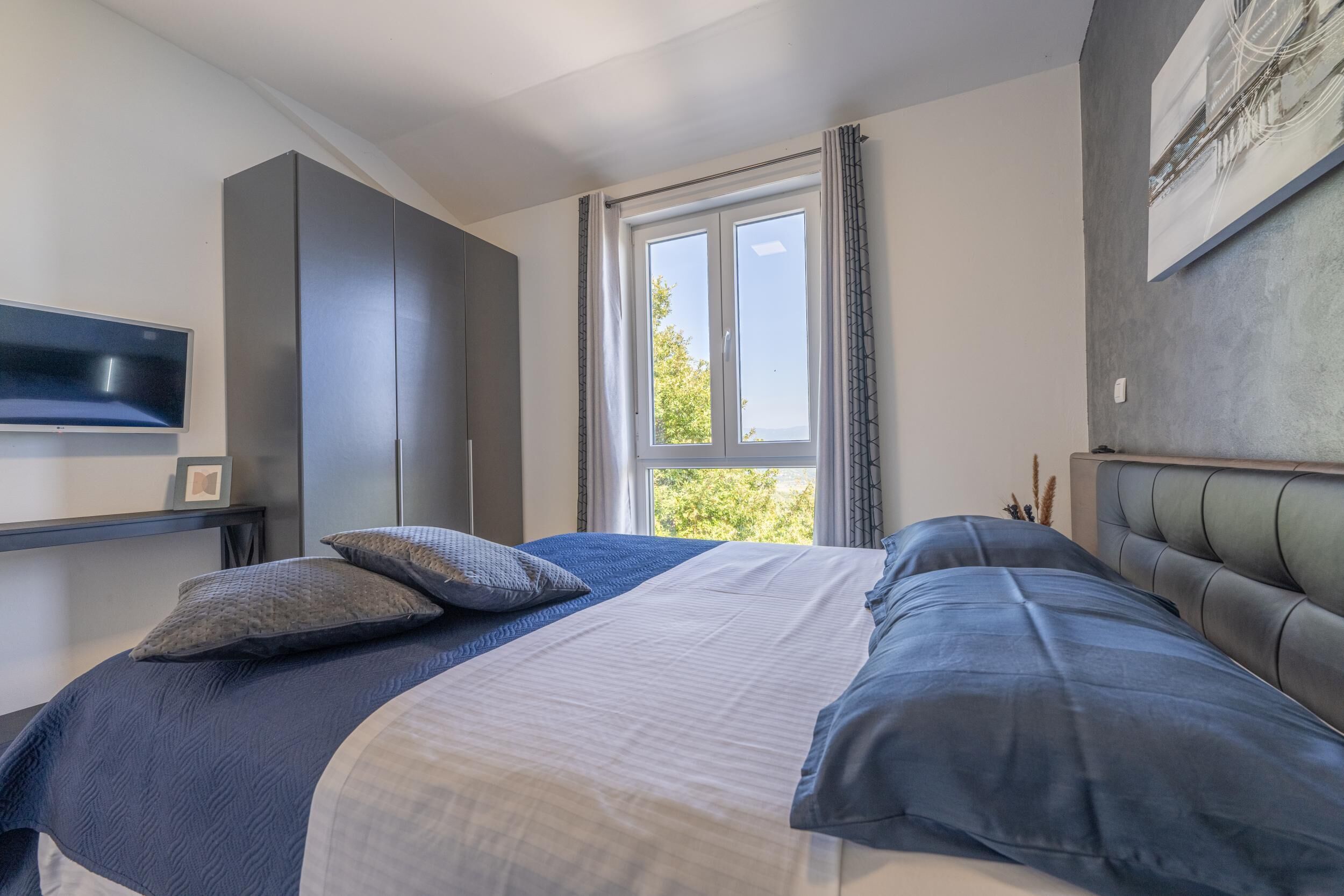 Ein Schlafzimmer mit einem großen Bett, einem Kleiderschrank, einem Flachbildfernseher und einem Fenster mit Vorhängen.