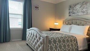 Een slaapkamer met een gestoffeerd hoofdeinde, een bed met een decoratief sprei, een bedlampje en een raam met gordijnen.