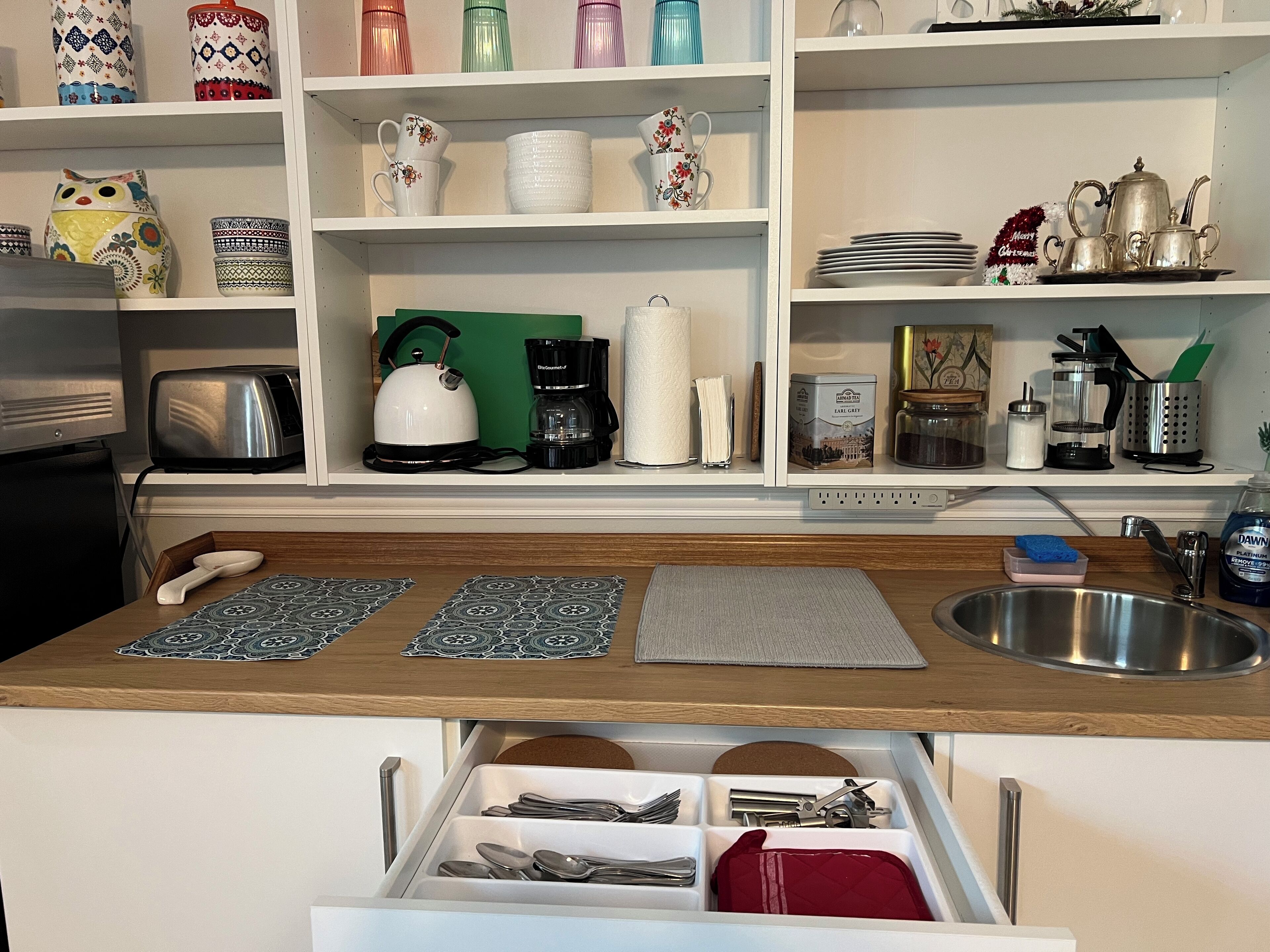 Una cocina con encimera de madera, un tostador, un hervidor, una cafetera y diferentes utensilios de cocina en estantes.
