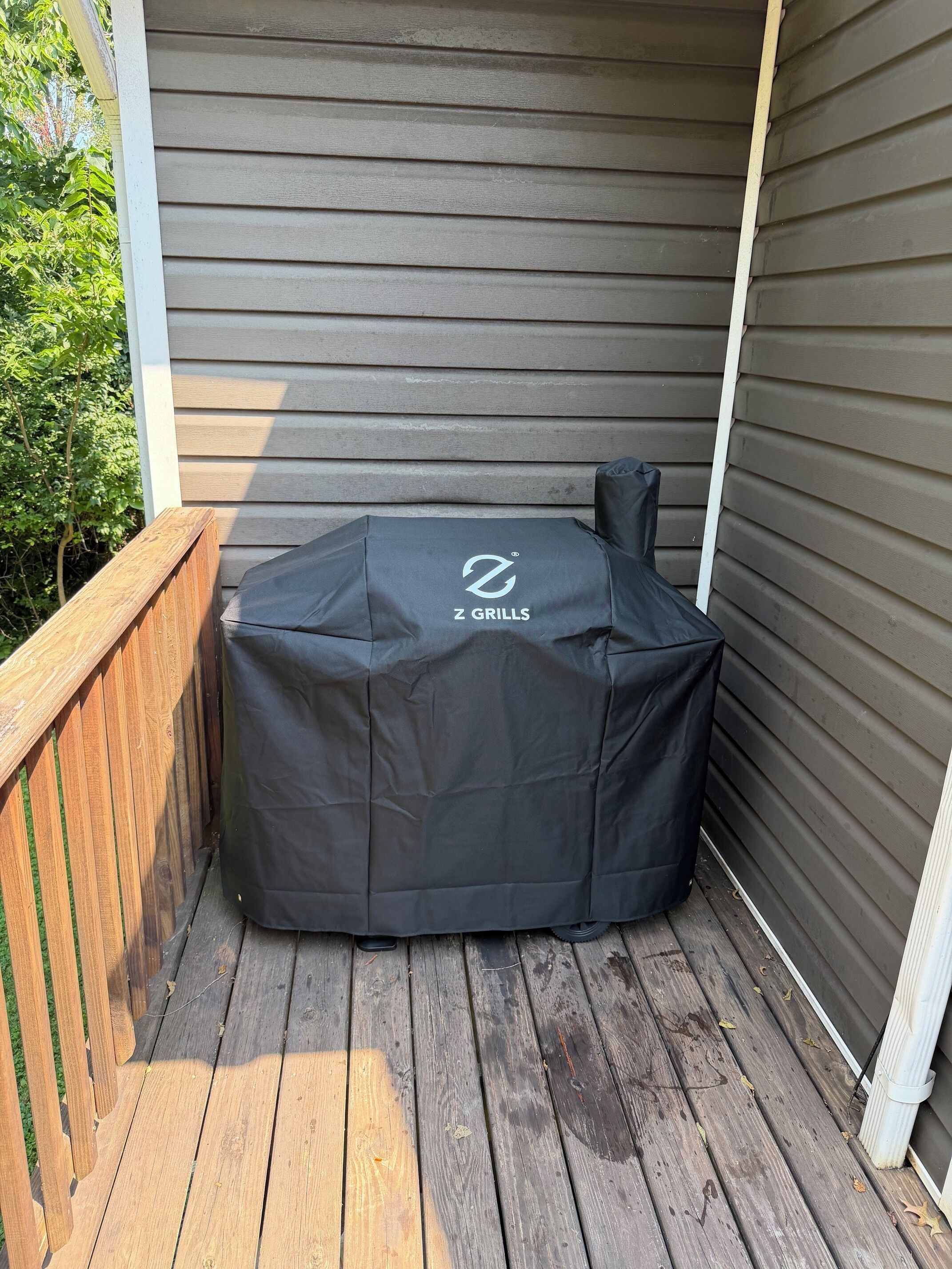 A covered grill with a logo, located on a wooden deck adjacent to a house with siding.