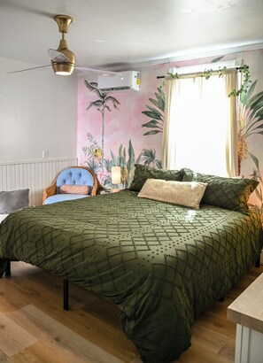 Quarto com cama, cadeira, mesa de cabeceira, ventilador de teto, ar condicionado e um mural tropical na parede.