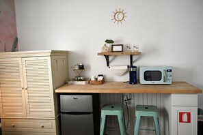 A compact kitchen with a microwave, a small refrigerator, a wooden table with a shelf, and two stools.