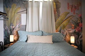 A bed with a quilted cover, two pillows, a nightstand with a lamp, and a wall with a tropical mural.