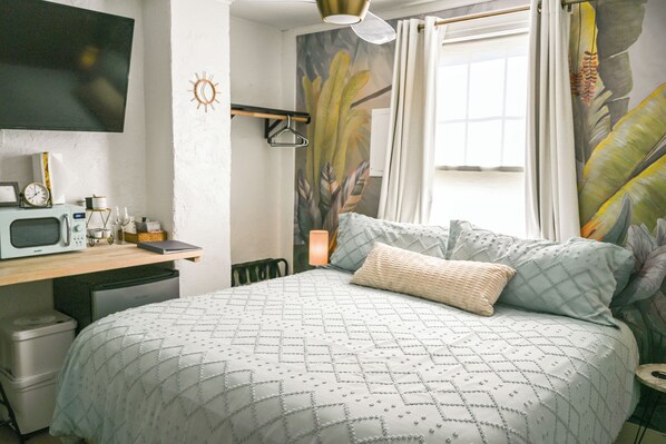 A bedroom with a bed, a desk with a microwave, a small fridge, a hanging light, and a window with curtains.