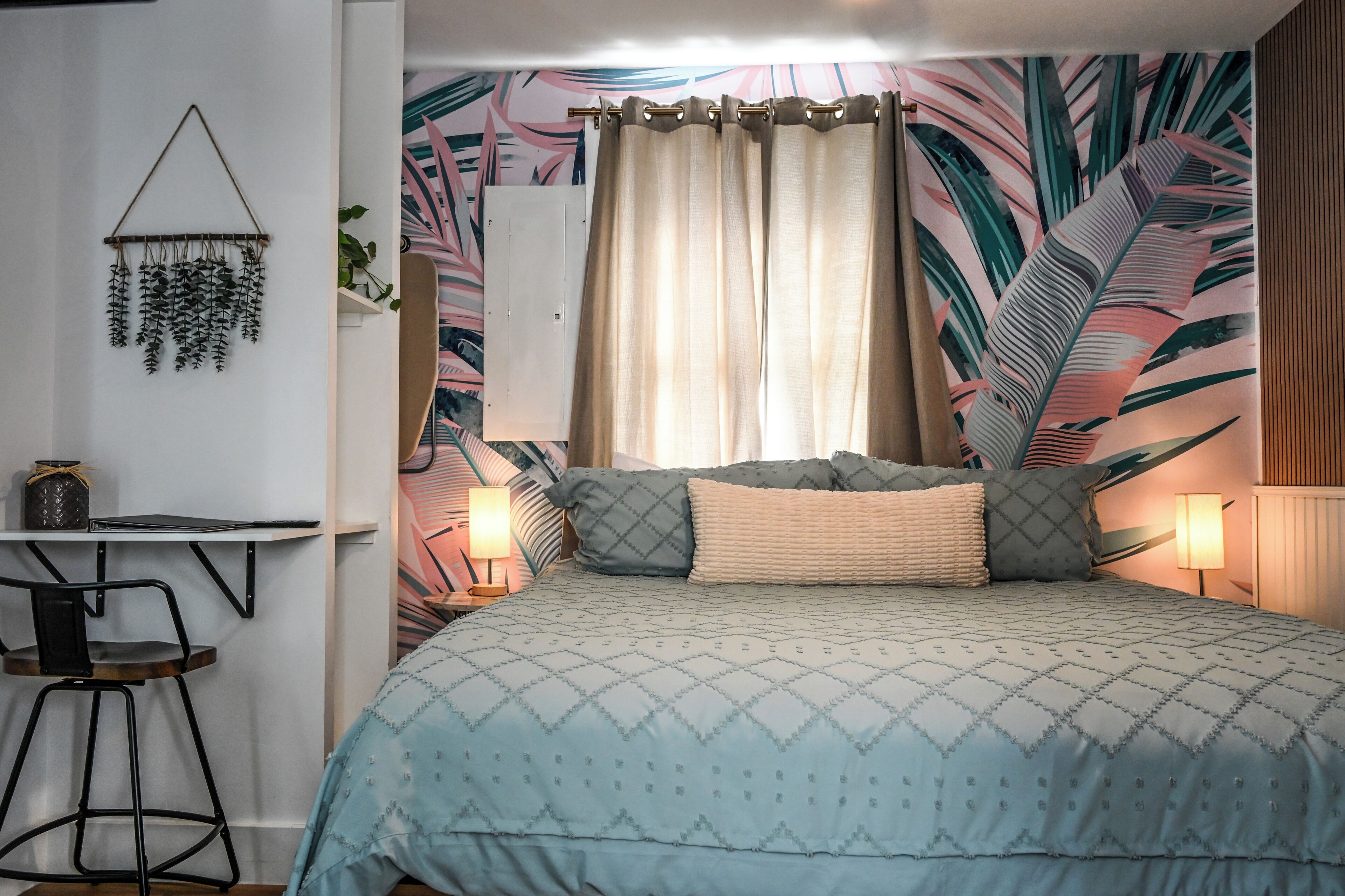 A bedroom with a bed, a desk, a chair, and a wall with a tropical leaf pattern.