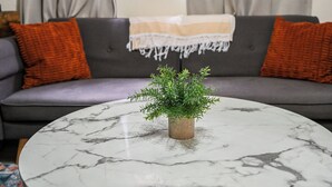 Ein Wohnzimmer mit grauem Sofa, orangefarbenen Kissen, einem weißen Marmor-Couchtisch und einer kleinen Topfpflanze.