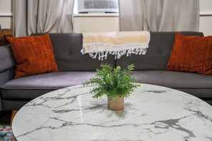 A living room with a grey sofa, orange cushions, a white marble coffee table, and a small potted plant.