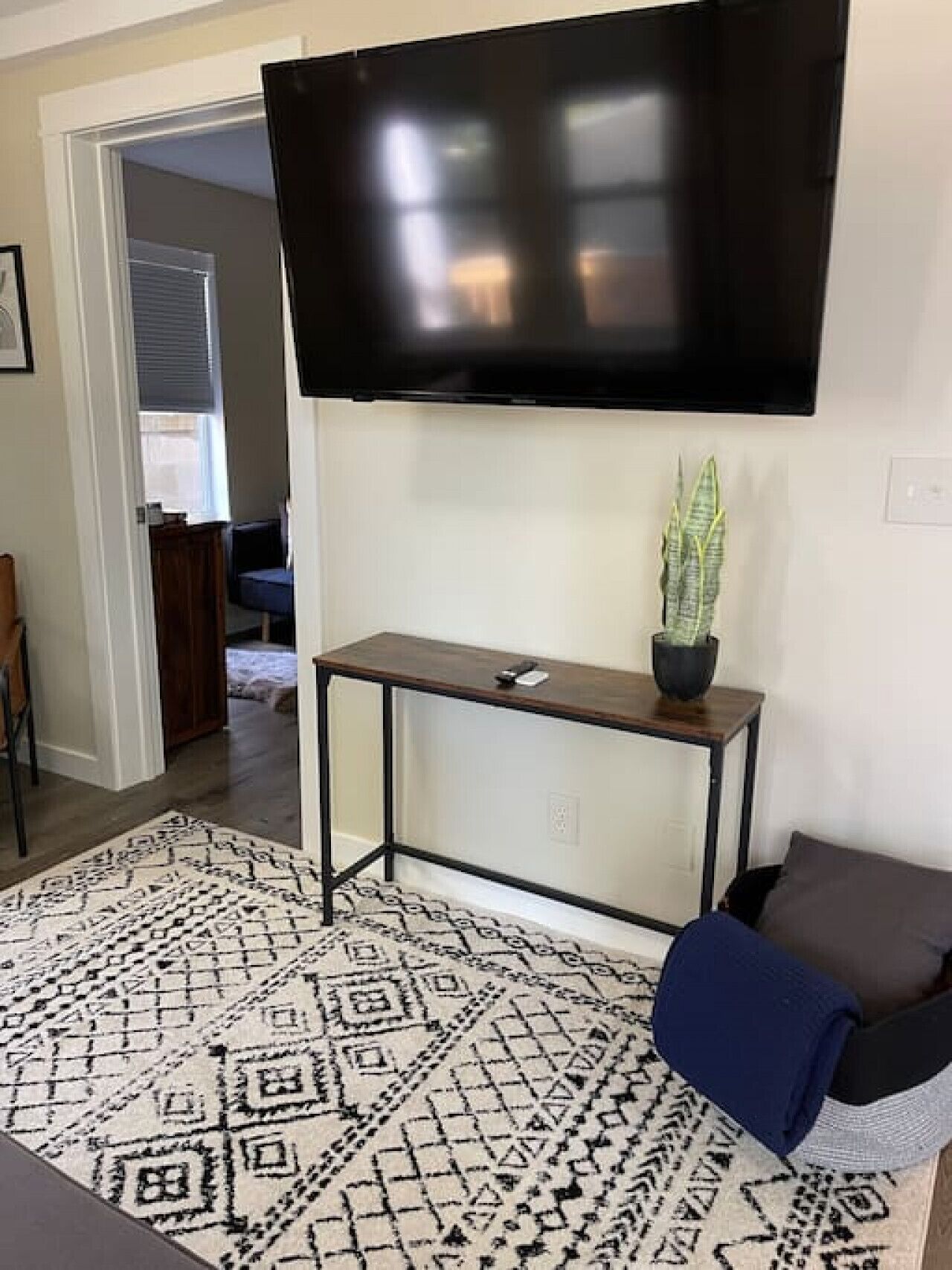 Un salon avec un téléviseur à écran plat fixé au mur, une petite table d’appoint en bois sur laquelle est posée une plante, un tapis à motifs et un canapé bleu.