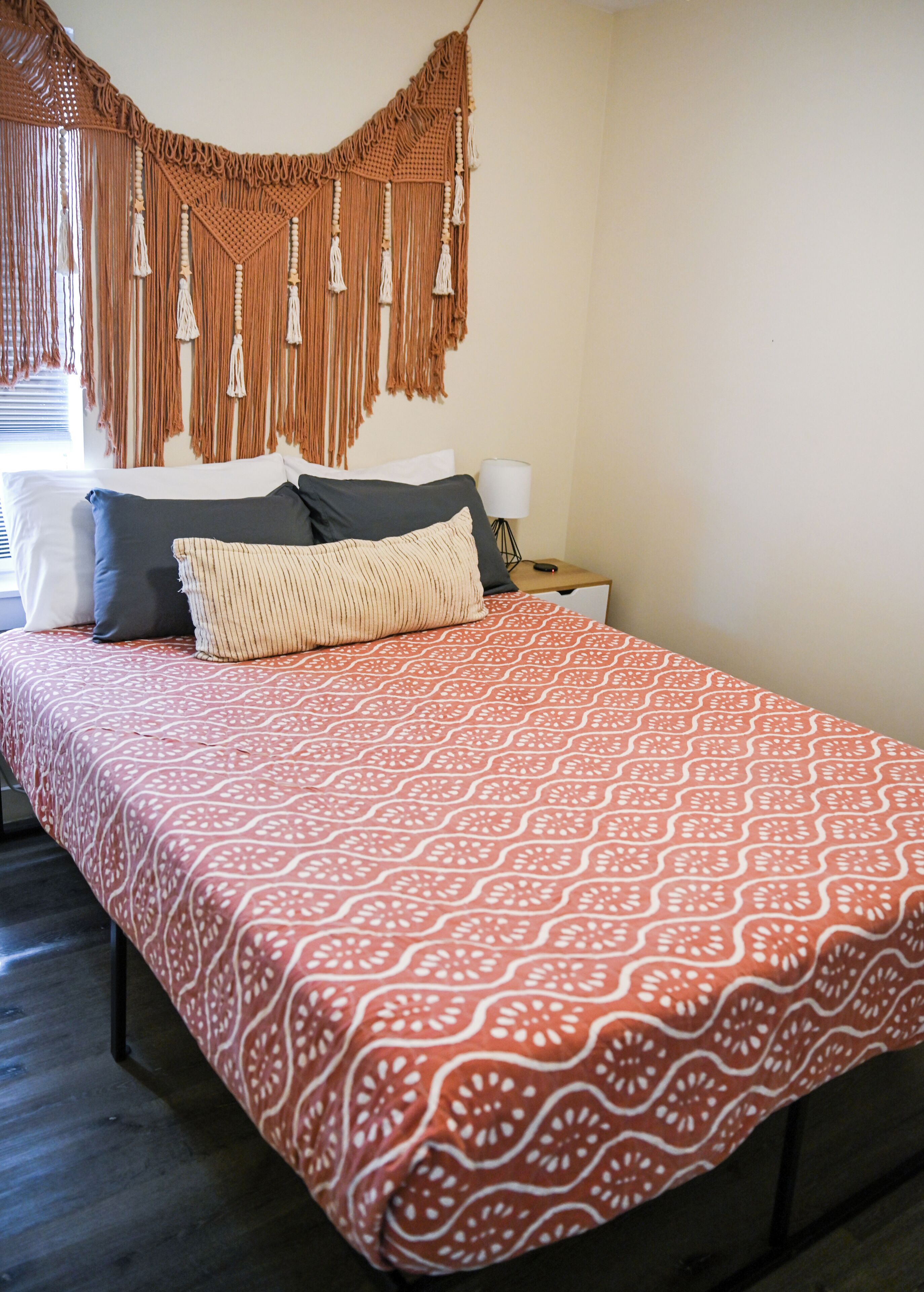 A bed with a patterned bedspread, a macrame wall hanging, and a bedside lamp.