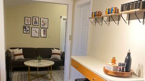 Una cocina compacta con encimera de madera, dos taburetes y una mesa de comedor. En el área de estar contigua, hay un sofá y una alfombra.