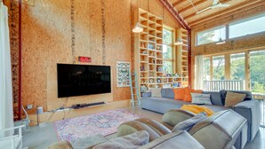 Un ampio soggiorno con una grande TV a schermo piatto montata a parete, un divano grigio, una libreria in legno e una finestra che offre vista sull'esterno.