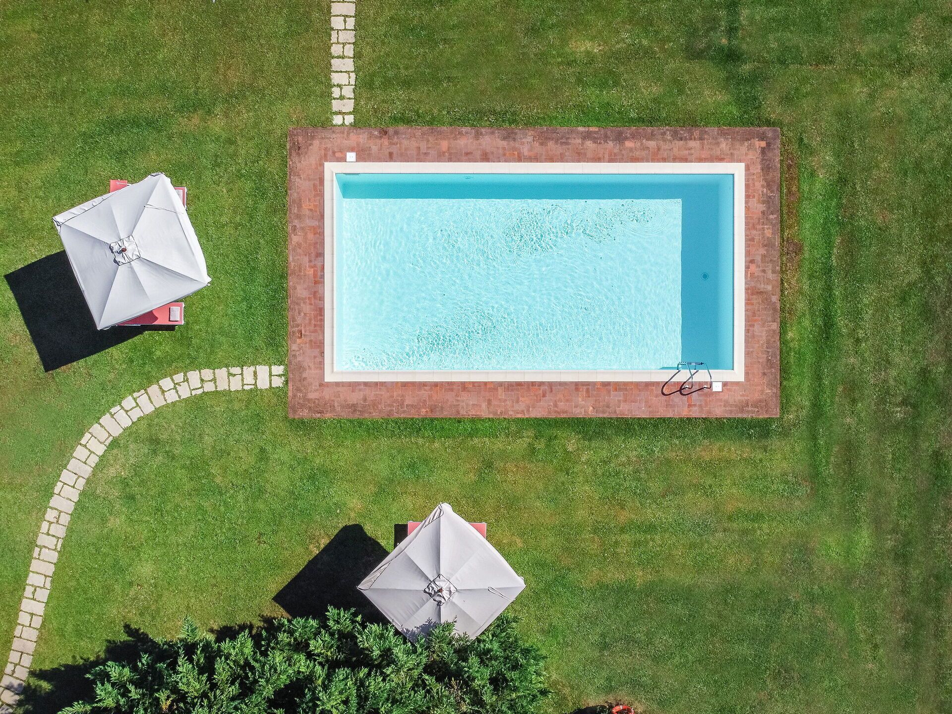 Vista aérea de una piscina rectangular con borde de ladrillo, rodeada por un césped cuidado y dos sombrillas.