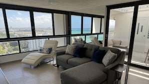 Een moderne woonkamer met een grijze modulaire bank, een witte fauteuil en een glazen salontafel. Grote ramen bieden uitzicht op de zee en de skyline.