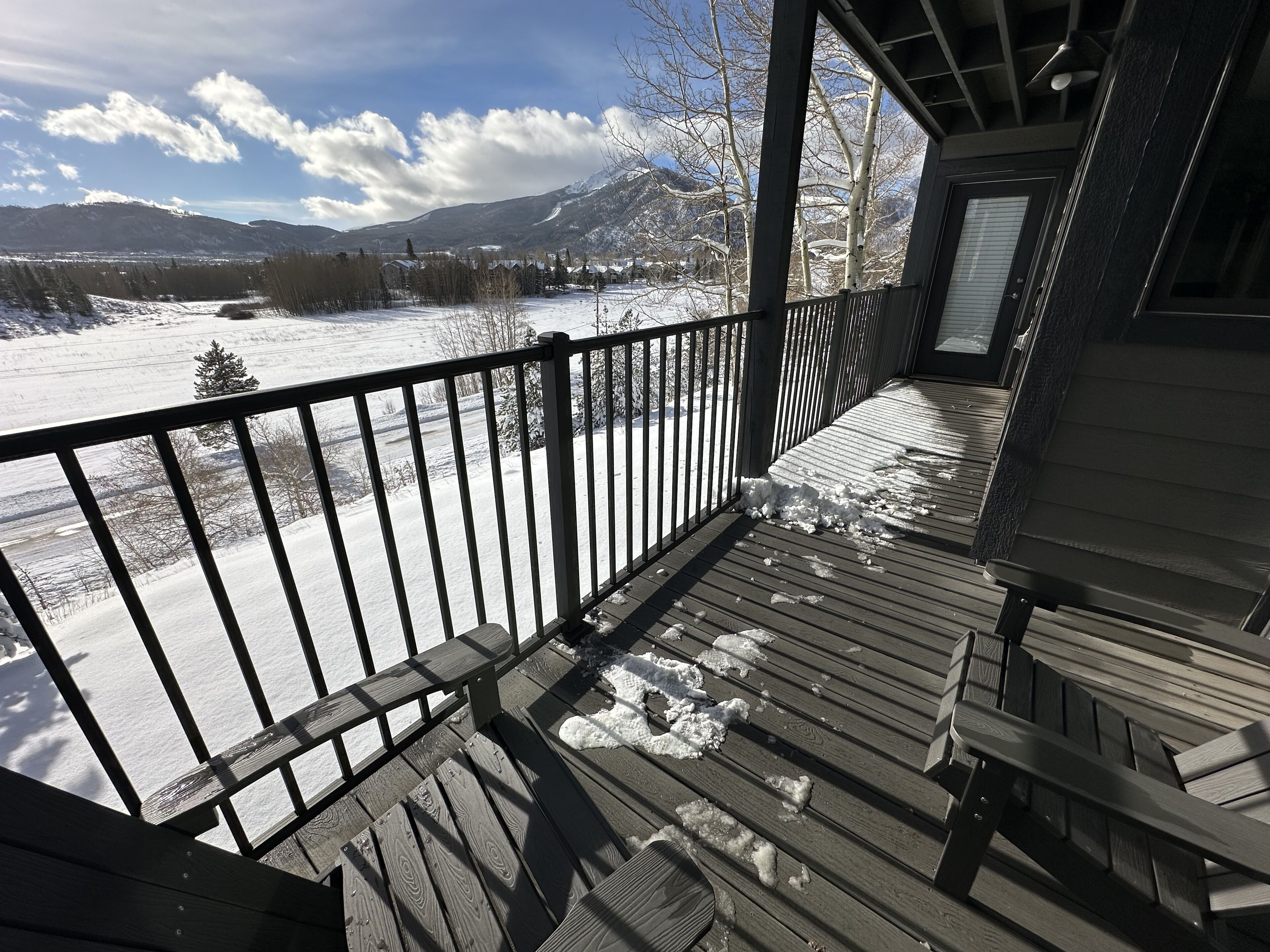 A wooden deck with snow on the ground and a view of a snowy landscape with mountains in the background.