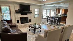 A spacious living room with a fireplace, leather sofa, and a dining area with a kitchen in the background.