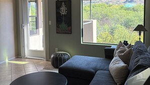 Uma sala de estar com sofá, uma mesa redonda, um ventilador de teto, uma janela grande com vista para as montanhas e uma porta que dá para o exterior.