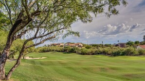 Um campo de golfe com uma árvore grande em primeiro plano, casas ao fundo e um céu claro.