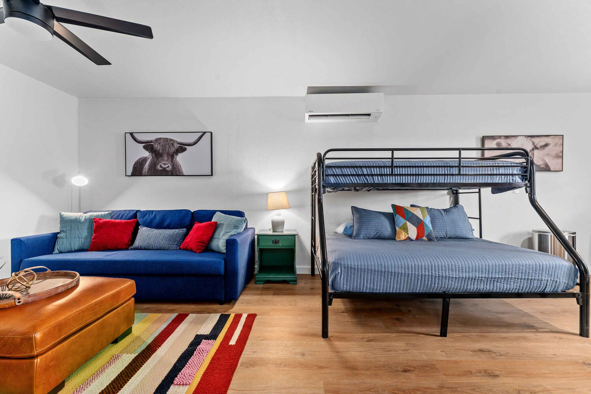 A room with a bunk bed, a blue sofa, a colorful striped rug, and a brown leather ottoman.