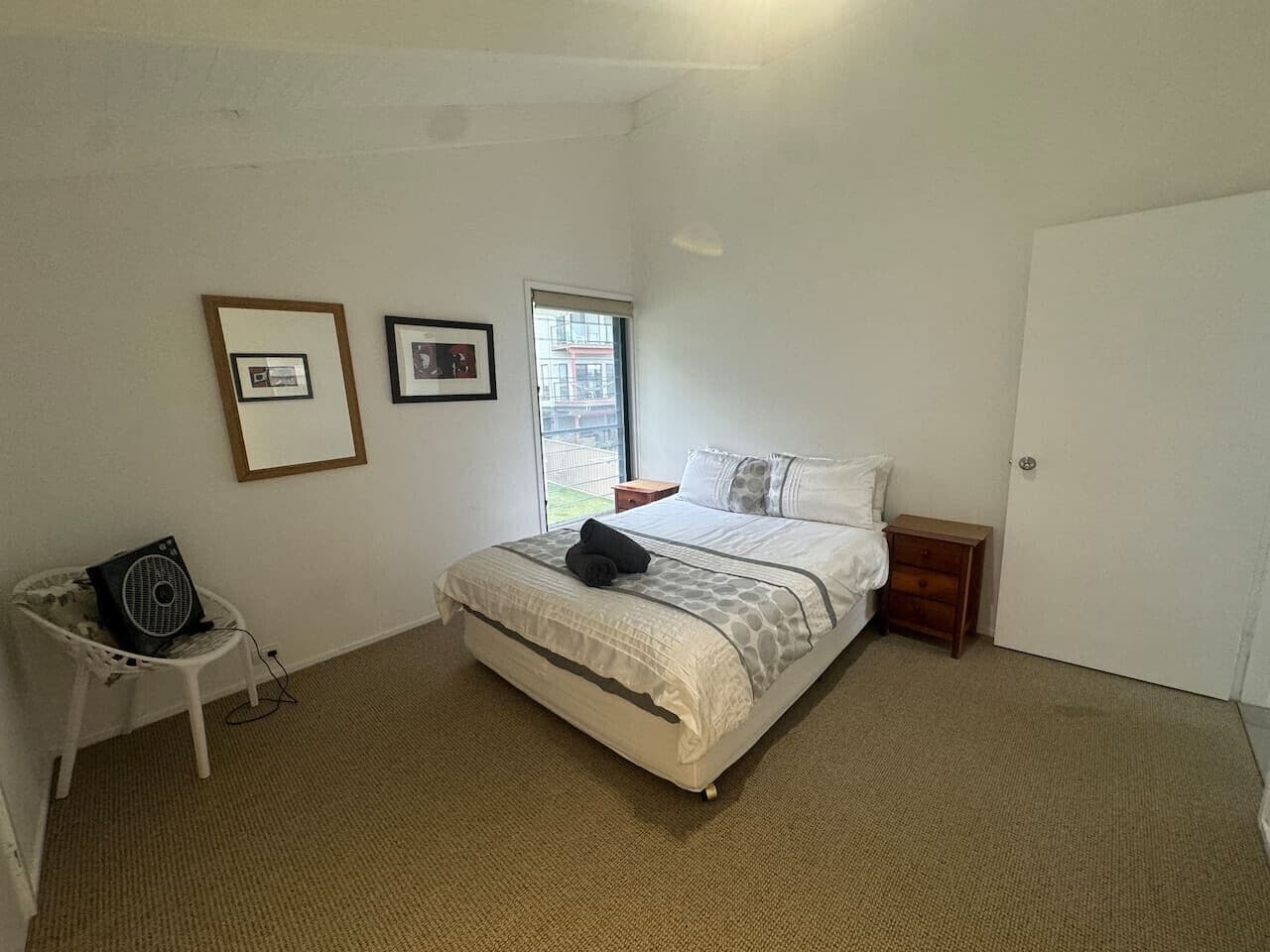A bedroom with a bed, a bedside table, a chair, a fan, and a window.