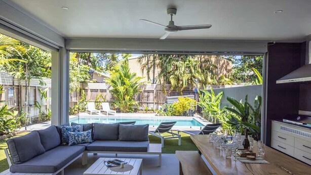 A modern outdoor living area with a pool, a sofa, a dining table, and a ceiling fan.