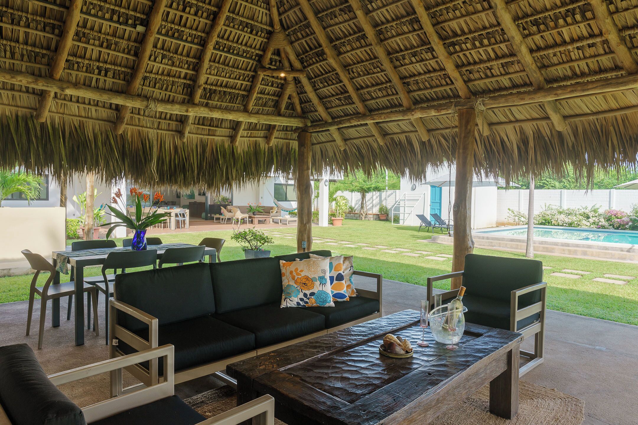 An outdoor seating area with a thatched roof, green cushioned sofas, and a wooden table.