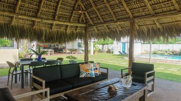An outdoor seating area with a thatched roof, green cushioned sofas, and a wooden table.