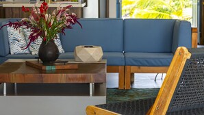 A modern living room with a blue sofa, a wooden coffee table with a vase of flowers, and a view of a kitchen and palm trees through a glass door.