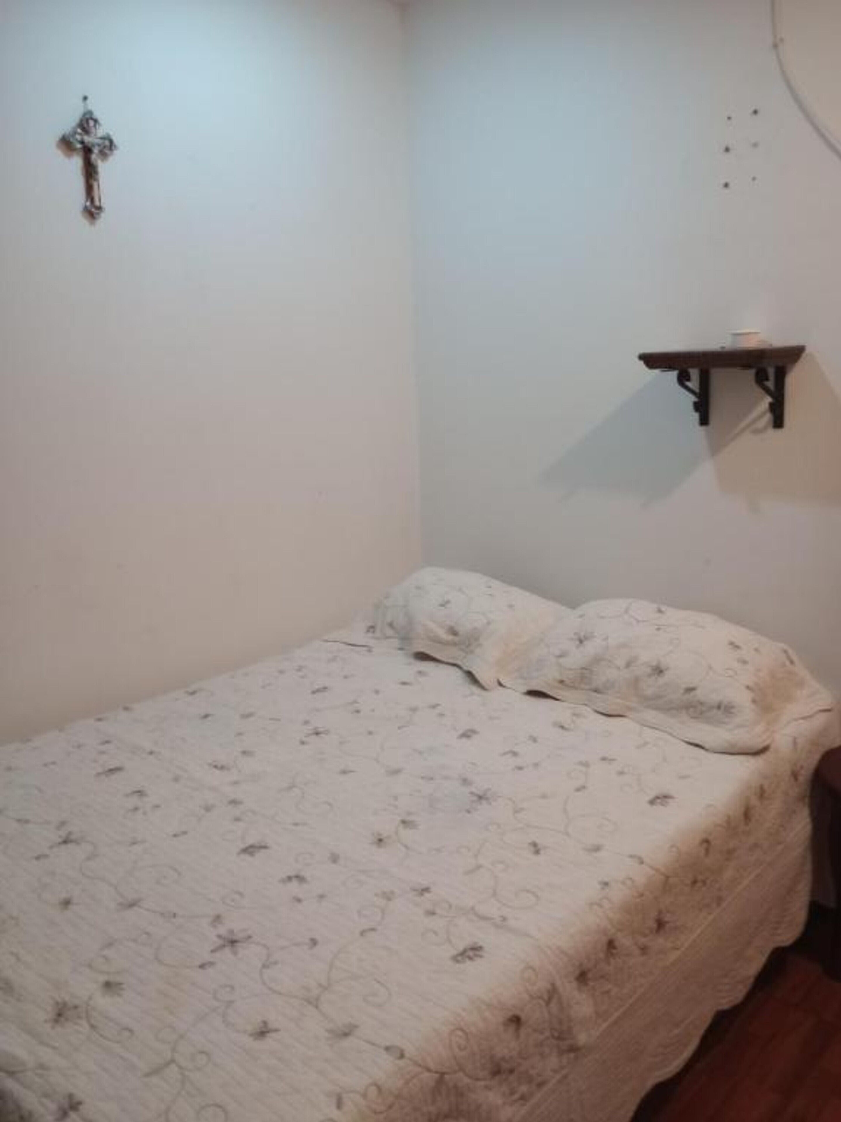 A small, simple bedroom with a single bed, a wall-mounted shelf, and a cross on the wall.