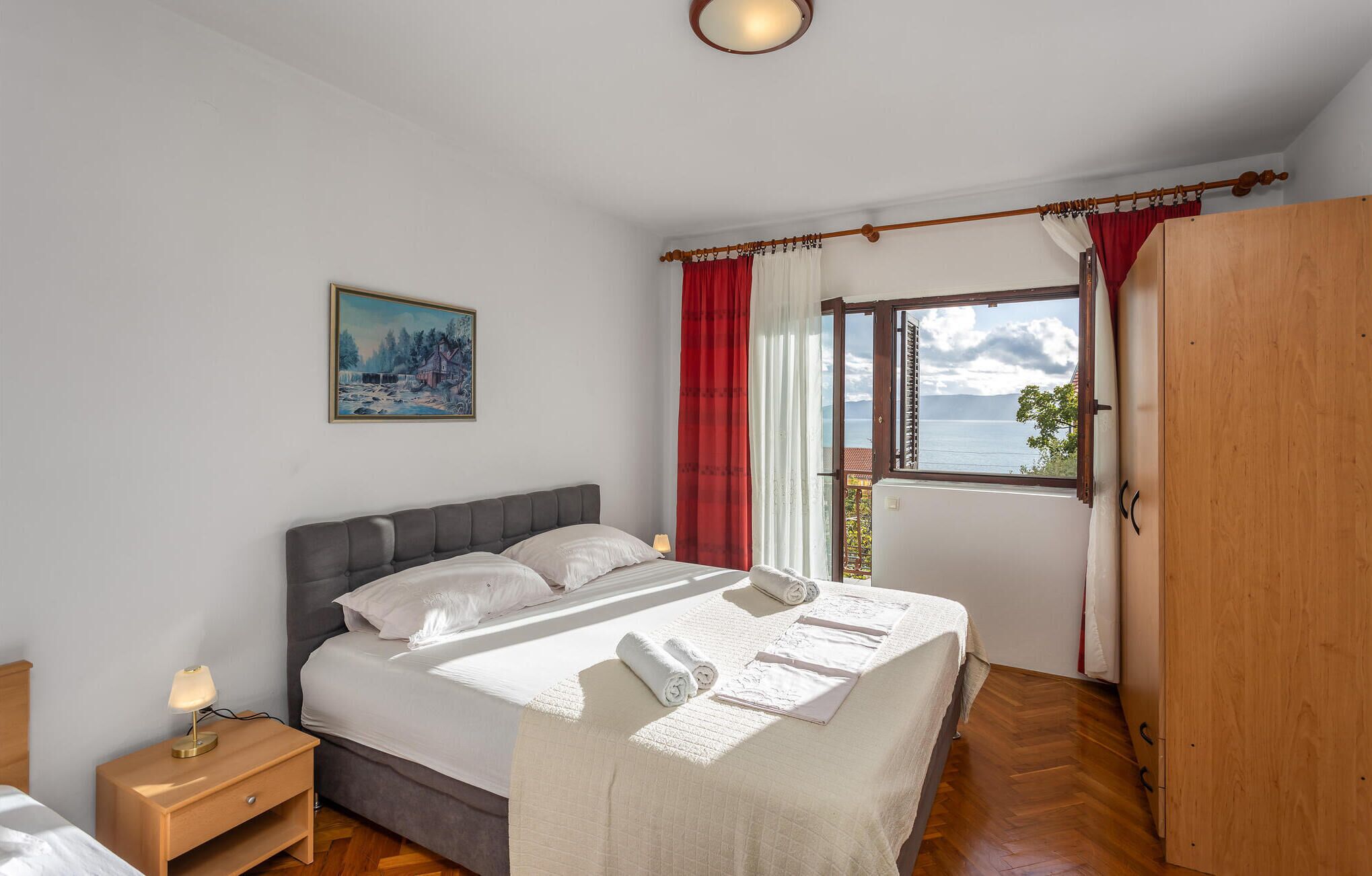 A bedroom with a bed, a nightstand, a painting, a window with curtains, and a view of the sea.