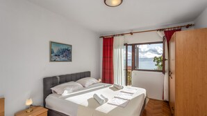 A bedroom with a bed, a nightstand, a painting, a window with curtains, and a view of the sea.